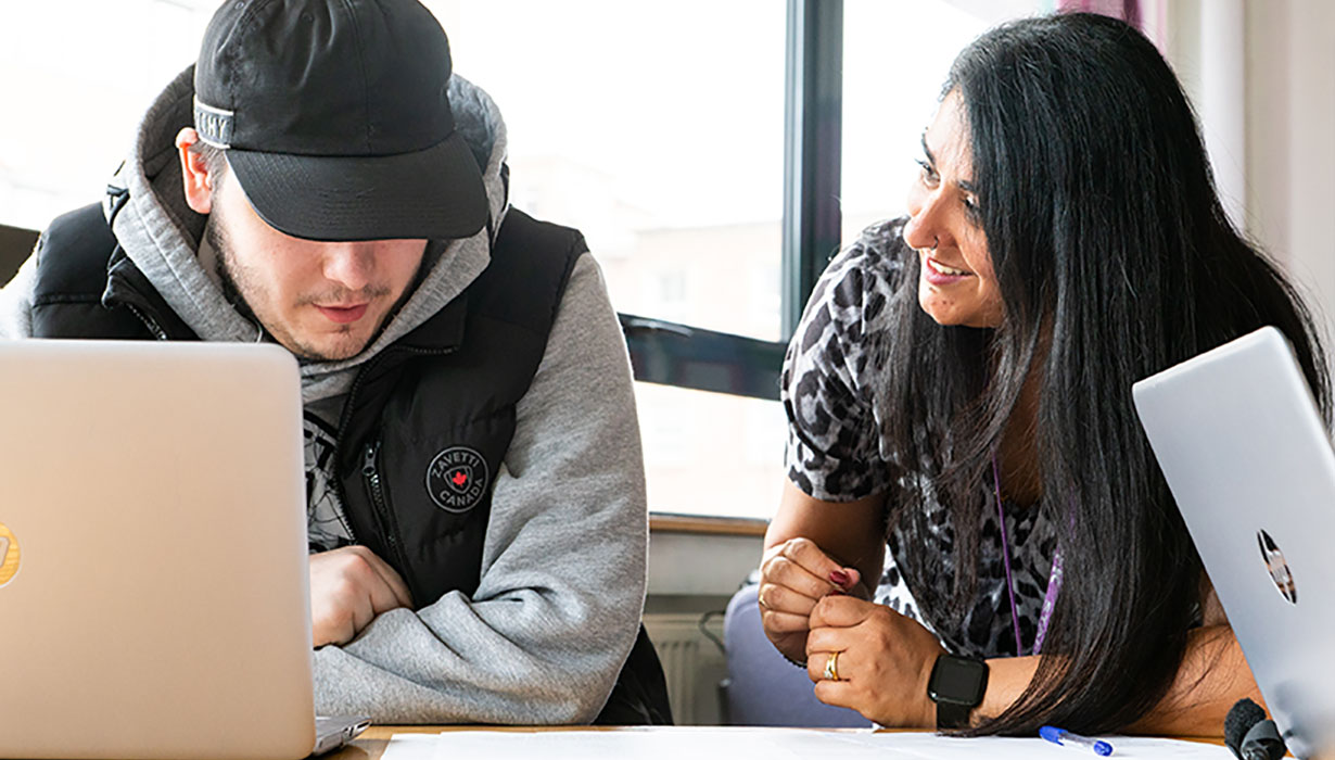 Careers adviser talking and looking at a young person who is sat at a desk working on a laptop in an office location